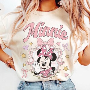 May include: An ivory t-shirt featuring a graphic of Minnie Mouse in a pink polka-dot bow, framed by ribbons and stars. The word Minnie is printed in large pink script above the character. The design includes small pink bows and yellow stars.