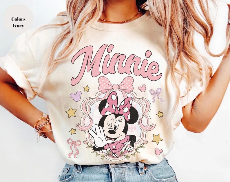 May include: An ivory t-shirt featuring a graphic of Minnie Mouse in a pink polka-dot bow, framed by ribbons and stars. The word Minnie is printed in large pink script above the character. The design includes small pink bows and yellow stars.