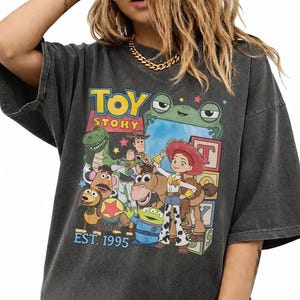 Può includere: T-shirt grigio scuro con una grafica colorata dei personaggi del film d'animazione "Toy Story". Il design include il testo "TOY STORY" e "EST. 1995."