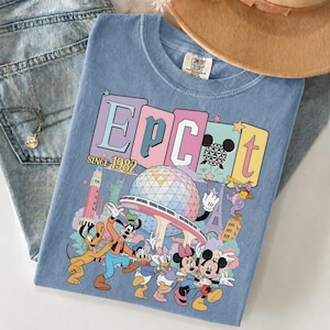 May include: A light blue Comfort Colors t-shirt featuring a colorful graphic of Disney characters and the text "EPCOT Since 1982". The design includes Mickey and Minnie Mouse, Goofy, Donald and Daisy Duck, and Pluto, with iconic landmarks.