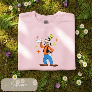 Puede incluir: Una sudadera rosa claro con una imagen bordada de Goofy, un personaje de dibujos animados de Disney. Goofy lleva una camisa naranja, pantalones azules y un sombrero verde. El dise&ntilde;o incluye estrellas naranjas y verdes. La palabra "Embroidered" est&aacute; en la parte inferior.