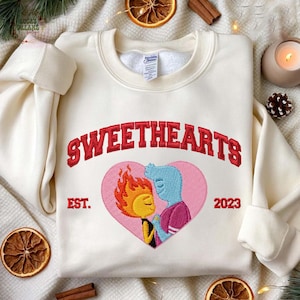 Op de afbeelding: Crèmekleurige sweatshirt met het woord "SWEETHEARTS" in rode, gebogen letters. Daaronder bevindt zich een roze hart met een geborduurd beeld van twee stripfiguren. De sweatshirt heeft ook "EST. 2023".