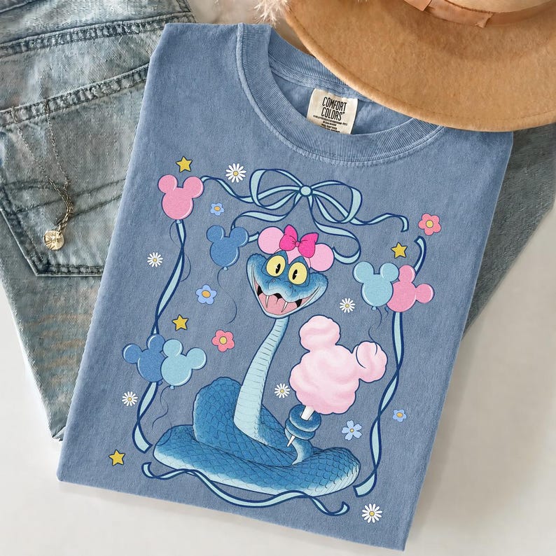 May include: A light blue Comfort Colors t-shirt featuring a cartoon snake holding cotton candy. The snake wears a pink bow and Mickey Mouse ears. The shirt is decorated with Mickey Mouse balloons, flowers, and stars.