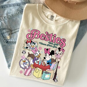 May include: Cream-colored t-shirt featuring the word "Besties" in pink script, with cartoon characters and the phrase "Girls Just Wanna Have Fun." The shirt is folded on top of blue jeans, with a necklace and a hat visible.