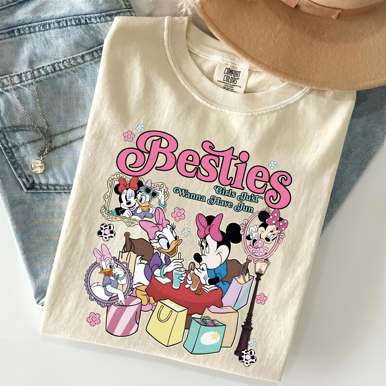 May include: Cream-colored t-shirt featuring the word "Besties" in pink script, with cartoon characters and the phrase "Girls Just Wanna Have Fun." The shirt is folded on top of blue jeans, with a necklace and a hat visible.