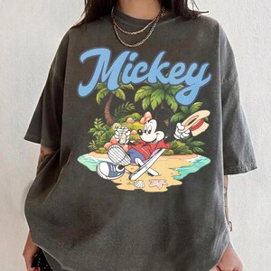 May include: Dark gray t-shirt featuring the word "Mickey" in blue script above a cartoon scene of Mickey Mouse relaxing on a beach. Mickey is wearing a red shirt and holding a drink and a hat. Palm trees and ocean waves are in the background.