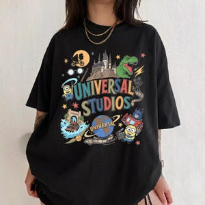 May include: Black t-shirt with a colorful graphic featuring the words "UNIVERSAL STUDIOS" and various theme park characters and imagery, including a dinosaur, Minions, and a castle.