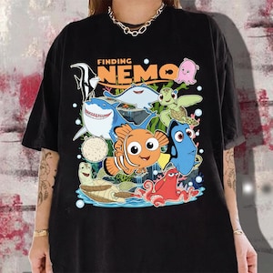 Op de afbeelding: Zwart T-shirt met een kleurrijke afbeelding van personages uit de film "Finding Nemo". Het ontwerp bevat Nemo, Dory, een haai, een schildpad en andere zeedieren. De woorden "Finding Nemo" staan boven de personages gedrukt.