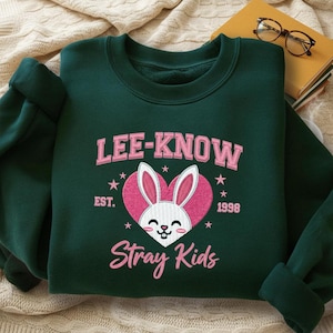 Könnte beinhalten: Ein dunkelgrünes Sweatshirt mit dem Text "LEE-KNOW" über einem rosa herzförmigen Hasen-Design. Unter dem Hasen steht der Text "Stray Kids". Das Sweatshirt hat auch den Text "EST. 1998" und mehrere Sterne.