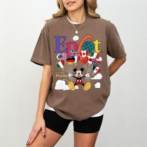 K&ouml;nnte beinhalten: Braunes T-Shirt mit einer farbenfrohen Grafik mit dem Wort "Epcot" und Mickey Mouse, die herzf&ouml;rmige Ballons mit Flaggen h&auml;lt. Das Shirt hat auch den Text "Est. 1982 Florida".