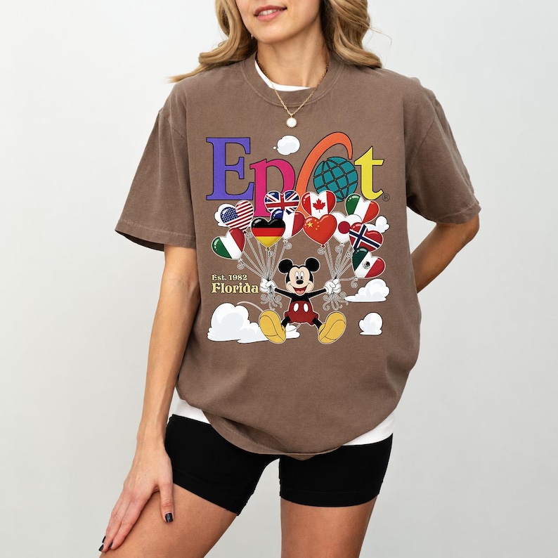 K&ouml;nnte beinhalten: Braunes T-Shirt mit einer farbenfrohen Grafik mit dem Wort "Epcot" und Mickey Mouse, die herzf&ouml;rmige Ballons mit Flaggen h&auml;lt. Das Shirt hat auch den Text "Est. 1982 Florida".