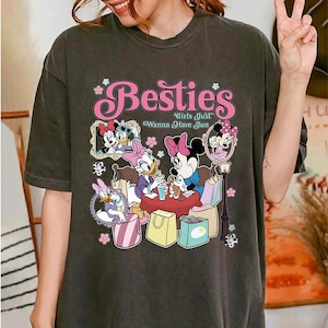 May include: Dark gray t-shirt featuring the word "Besties" in pink script with floral accents. The shirt showcases a graphic of Disney characters Minnie Mouse, Daisy Duck, and others, with the phrase "Girls Just Wanna Have Jun".