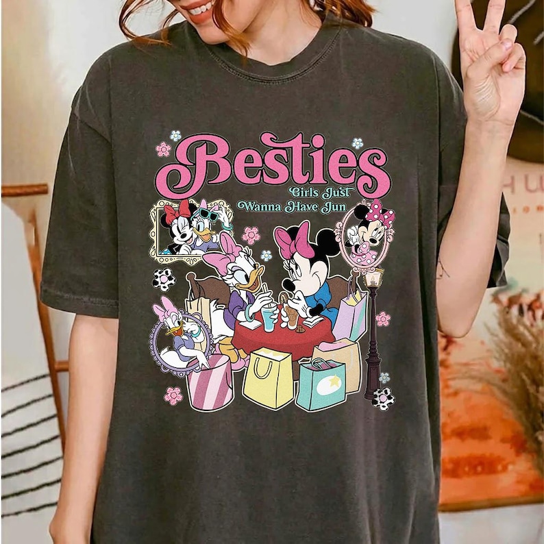 May include: Dark gray t-shirt featuring the word "Besties" in pink script with floral accents. The shirt showcases a graphic of Disney characters Minnie Mouse, Daisy Duck, and others, with the phrase "Girls Just Wanna Have Jun".