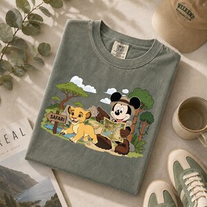 Op de afbeelding: Saliegroen t-shirt met een afbeelding van Mickey Mouse en Simba uit The Lion King op safari. Mickey draagt een safarihoed en houdt een kaart vast. Simba zit naast een bord met "Safari".