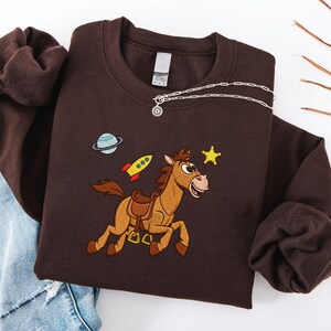 Pu&ograve; includere: Una felpa marrone scuro con un design ricamato raffigurante un cavallo dei cartoni animati, un pianeta, un razzo e una stella. La felpa ha un girocollo e maniche lunghe. Una collana d'argento con un piccolo ciondolo &egrave; drappeggiata sul davanti.