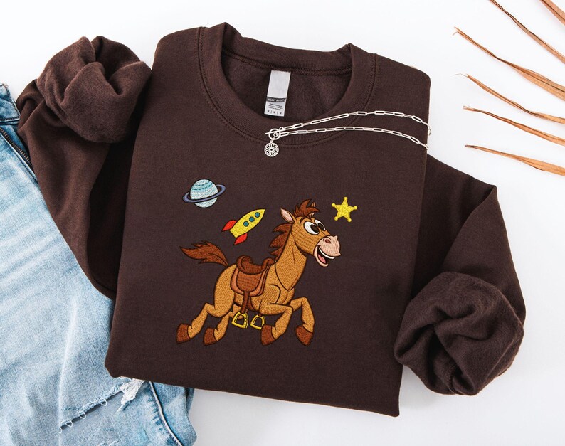 Pu&ograve; includere: Una felpa marrone scuro con un design ricamato raffigurante un cavallo dei cartoni animati, un pianeta, un razzo e una stella. La felpa ha un girocollo e maniche lunghe. Una collana d'argento con un piccolo ciondolo &egrave; drappeggiata sul davanti.