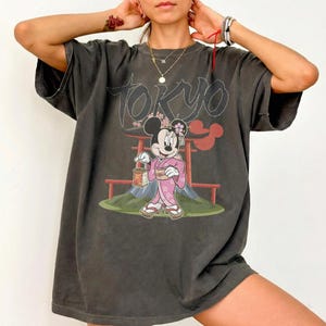Peut inclure: T-shirt gris fonc&eacute; surdimensionn&eacute; avec un motif de Minnie Mouse en kimono tenant une lanterne devant une porte torii japonaise et le Mont Fuji. Le mot "TOKYO" est au-dessus du motif.