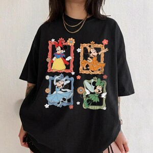 May include: Black t-shirt featuring four framed images of Minnie Mouse in different outfits. Each frame is decorated with flowers. Minnie Mouse is depicted in a yellow dress, orange dress, blue dress, and green fairy outfit.