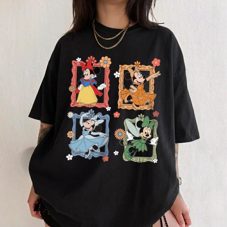 May include: Black t-shirt featuring four framed images of Minnie Mouse in different outfits. Each frame is decorated with flowers. Minnie Mouse is depicted in a yellow dress, orange dress, blue dress, and green fairy outfit.