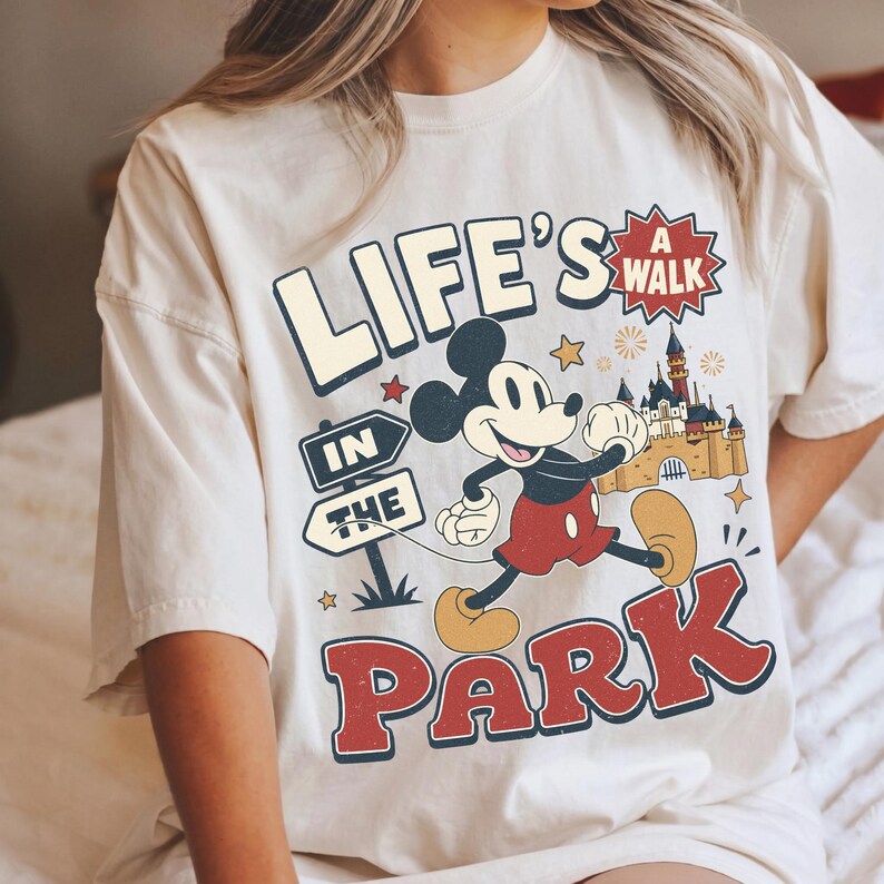 Puede incluir: Una camiseta color crema con un gr&aacute;fico de Mickey Mouse corriendo frente a un castillo. El texto dice "LIFE'S A WALK IN THE PARK" con "A WALK" dentro de una estrella roja. El dise&ntilde;o incluye se&ntilde;ales, peque&ntilde;as estrellas y fuegos artificiales.
