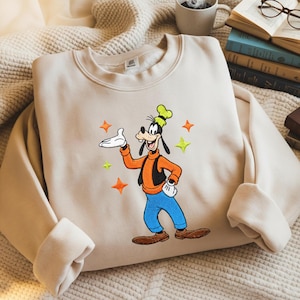 Puede incluir: Sudadera beige con una imagen bordada de Goofy, un personaje de dibujos animados, con su atuendo caracter&iacute;stico. Goofy lleva una camisa naranja, pantalones azules y un sombrero verde. El dise&ntilde;o incluye estrellas naranjas y verdes.