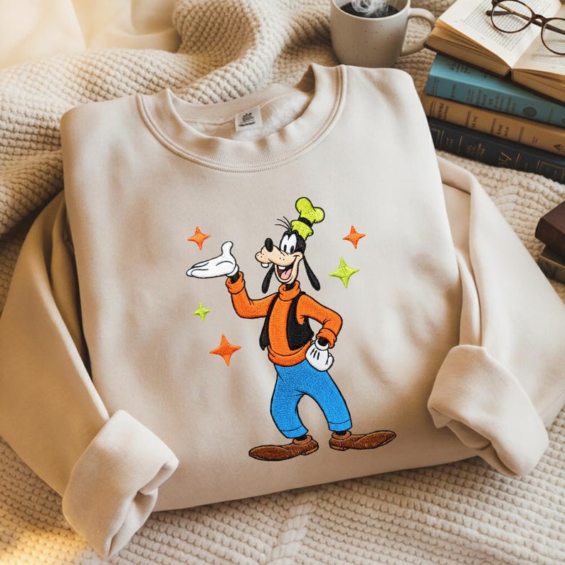 Puede incluir: Sudadera beige con una imagen bordada de Goofy, un personaje de dibujos animados, con su atuendo caracter&iacute;stico. Goofy lleva una camisa naranja, pantalones azules y un sombrero verde. El dise&ntilde;o incluye estrellas naranjas y verdes.