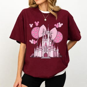 May include: Burgundy t-shirt featuring a pink castle graphic with a large bow and Mickey Mouse balloons. The castle is detailed with turrets and a central tower. The shirt has a white crew neck and short sleeves.