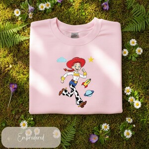 May include: A pale pink sweatshirt with an embroidered cartoon cowgirl design. The cowgirl is running, wearing a red hat, and is accompanied by a rocket and planet. The word "Embroidered" is at the bottom.
