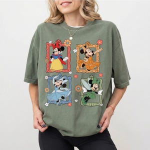 May include: Olive green t-shirt featuring four framed Minnie Mouse illustrations in different outfits. The illustrations are in red, orange, blue, and green frames with floral accents. The shirt has a relaxed fit.