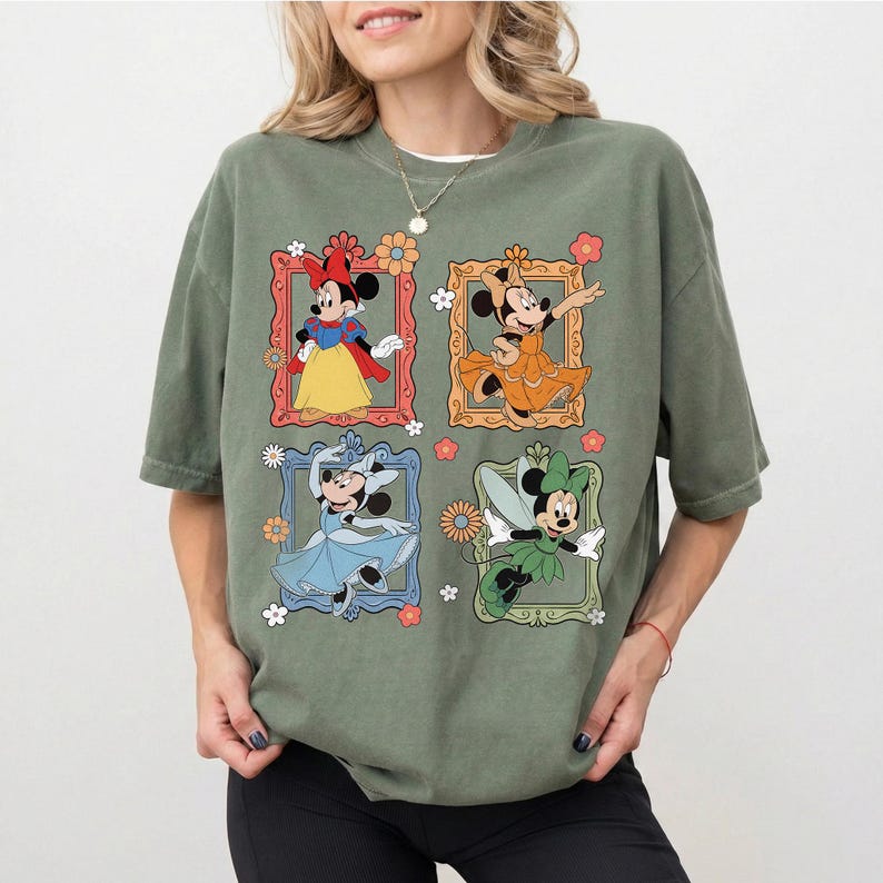 May include: Olive green t-shirt featuring four framed Minnie Mouse illustrations in different outfits. The illustrations are in red, orange, blue, and green frames with floral accents. The shirt has a relaxed fit.
