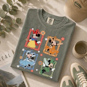 May include: A sage green t-shirt featuring four framed images of Minnie Mouse in different outfits. The frames are decorated with flowers. A Comfort Colors tag is visible at the top. A tan baseball cap, a mug, and sneakers are also in the image.