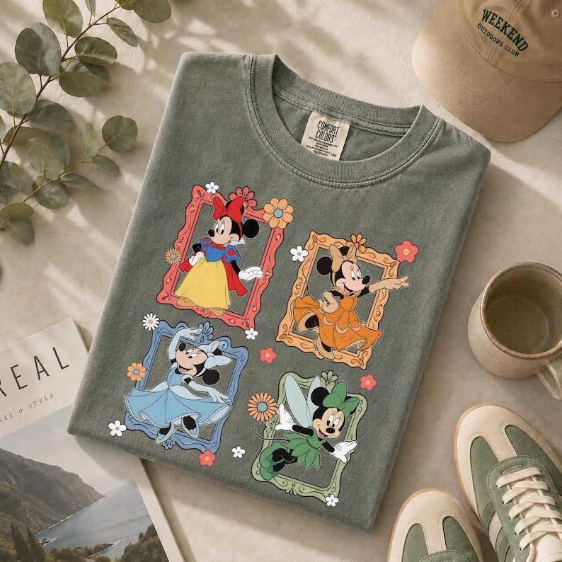 May include: A sage green t-shirt featuring four framed images of Minnie Mouse in different outfits. The frames are decorated with flowers. A Comfort Colors tag is visible at the top. A tan baseball cap, a mug, and sneakers are also in the image.