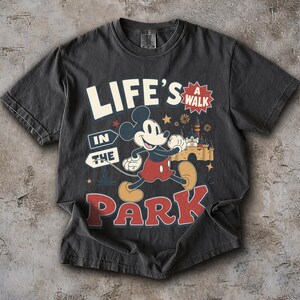 Puede incluir: Una camiseta gris carb&oacute;n con un gr&aacute;fico de Mickey Mouse caminando. El texto "LIFE'S A WALK IN THE PARK" aparece en letras blancas y rojas. El dise&ntilde;o incluye una ilustraci&oacute;n de un castillo, peque&ntilde;as estrellas y fuegos artificiales en amarillo y azul.