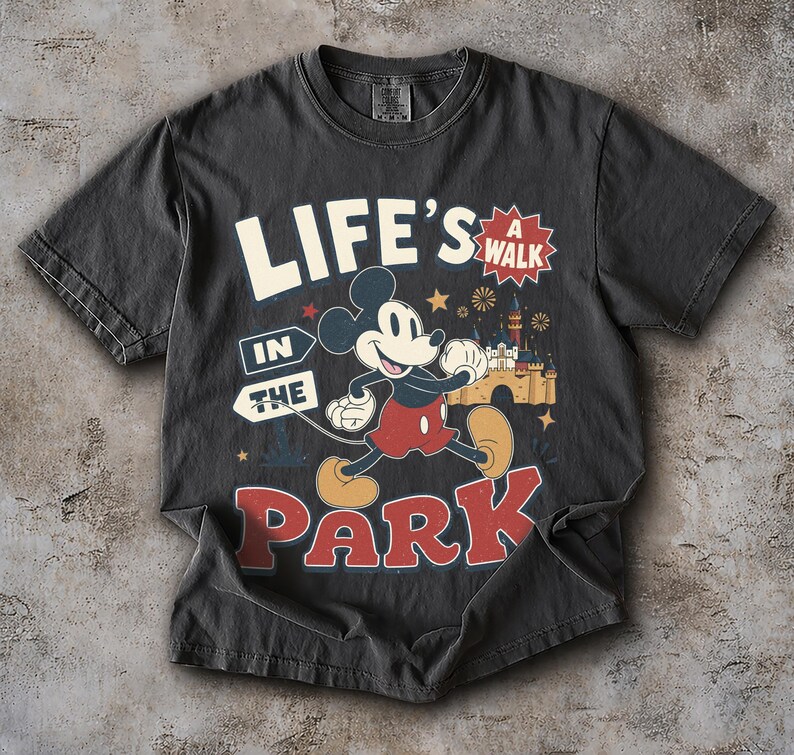 Puede incluir: Una camiseta gris carb&oacute;n con un gr&aacute;fico de Mickey Mouse caminando. El texto "LIFE'S A WALK IN THE PARK" aparece en letras blancas y rojas. El dise&ntilde;o incluye una ilustraci&oacute;n de un castillo, peque&ntilde;as estrellas y fuegos artificiales en amarillo y azul.