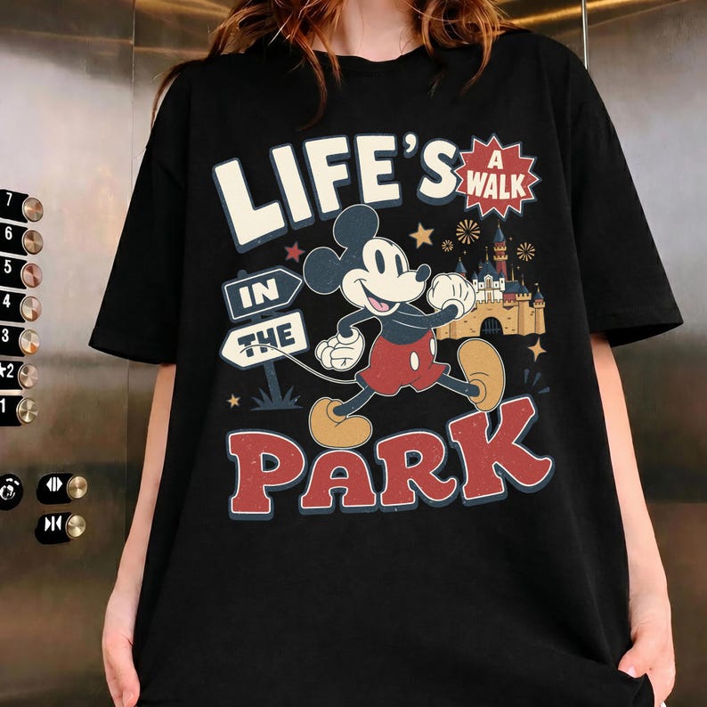 Puede incluir: Una camiseta negra de manga corta con un gr&aacute;fico de Mickey Mouse caminando. El texto dice "LIFE'S A WALK IN THE PARK". El dise&ntilde;o incluye un castillo, estrellas y fuegos artificiales en tonos rojo, amarillo, azul y blanco sobre la tela negra.