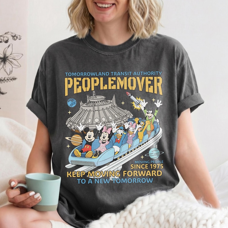 May include: Dark gray t-shirt featuring a Tomorrowland PeopleMover design with Mickey Mouse, Minnie Mouse, Donald Duck, and Goofy. The shirt includes the text "TOMORROWLAND TRANSIT AUTHORITY PEOPLEMOVER" and "KEEP MOVING FORWARD TO A NEW TOMORROW."