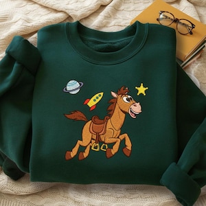 Pu&ograve; includere: Felpa verde scuro con un cavallo dei cartoni animati ricamato, una stella gialla, un razzo e un pianeta. Il cavallo &egrave; marrone con la bocca rosa e sta correndo. La felpa ha un girocollo e maniche lunghe.