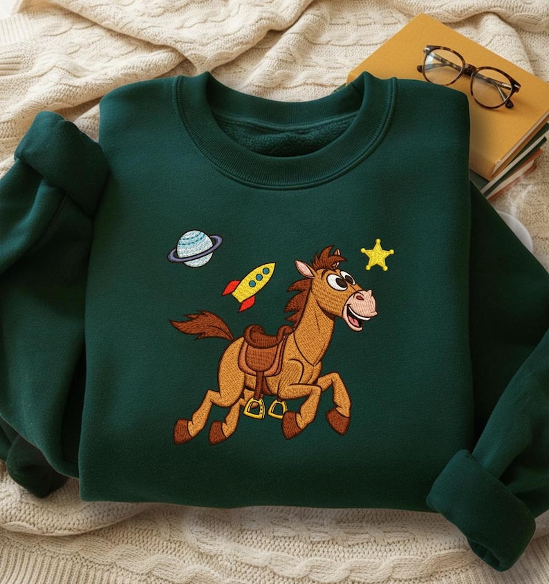 Pu&ograve; includere: Felpa verde scuro con un cavallo dei cartoni animati ricamato, una stella gialla, un razzo e un pianeta. Il cavallo &egrave; marrone con la bocca rosa e sta correndo. La felpa ha un girocollo e maniche lunghe.