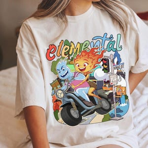 Puede incluir: Camiseta color crema con un gráfico colorido de dos personajes animados en una motocicleta. La palabra "Elemental" está escrita encima de los personajes en una fuente de arcoíris. Un letrero de la calle dice "Nomad St".