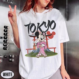 Peut inclure: T-shirt blanc avec un motif de Minnie Mouse en kimono rose tenant une lanterne, avec le mot "TOKYO" en noir au-dessus. Le motif comprend un torii rouge et un paysage de montagne.