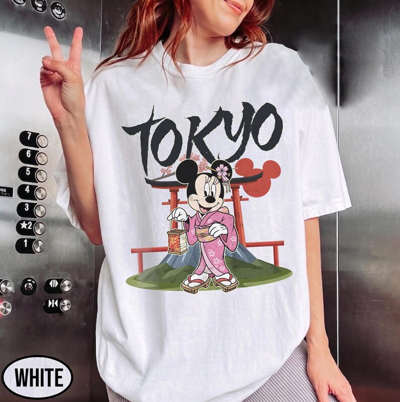Peut inclure: T-shirt blanc avec un motif de Minnie Mouse en kimono rose tenant une lanterne, avec le mot "TOKYO" en noir au-dessus. Le motif comprend un torii rouge et un paysage de montagne.