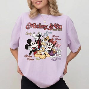 May include: A light purple t-shirt featuring a graphic of Mickey Mouse, Goofy, Donald Duck, Minnie Mouse, and Daisy Duck. The text "Mickey & Co Since 1928" is printed above the characters' names.