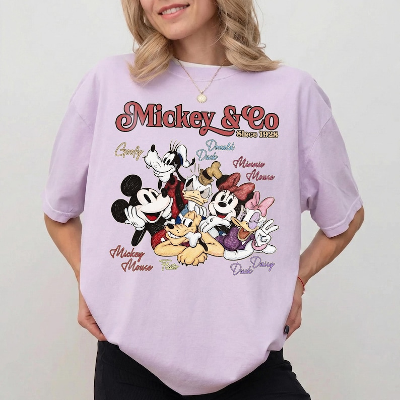 May include: A light purple t-shirt featuring a graphic of Mickey Mouse, Goofy, Donald Duck, Minnie Mouse, and Daisy Duck. The text "Mickey & Co Since 1928" is printed above the characters' names.
