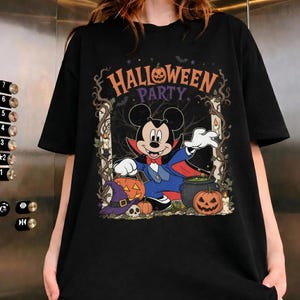 May include: A black t-shirt featuring a graphic of Mickey Mouse dressed as a vampire. The design includes the text "HALLOWEEN PARTY," pumpkins, a witch's hat, a skull, a cauldron, and lit candles framed by dark, twisted tree branches.