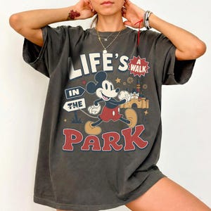 Puede incluir: Una camiseta gris carb&oacute;n de manga corta con un gr&aacute;fico de Mickey Mouse caminando. El texto dice "LIFE'S A WALK IN THE PARK" con una ilustraci&oacute;n de un castillo, estrellas y fuegos artificiales. El dise&ntilde;o usa una paleta de colores rojo, beige y blanco.