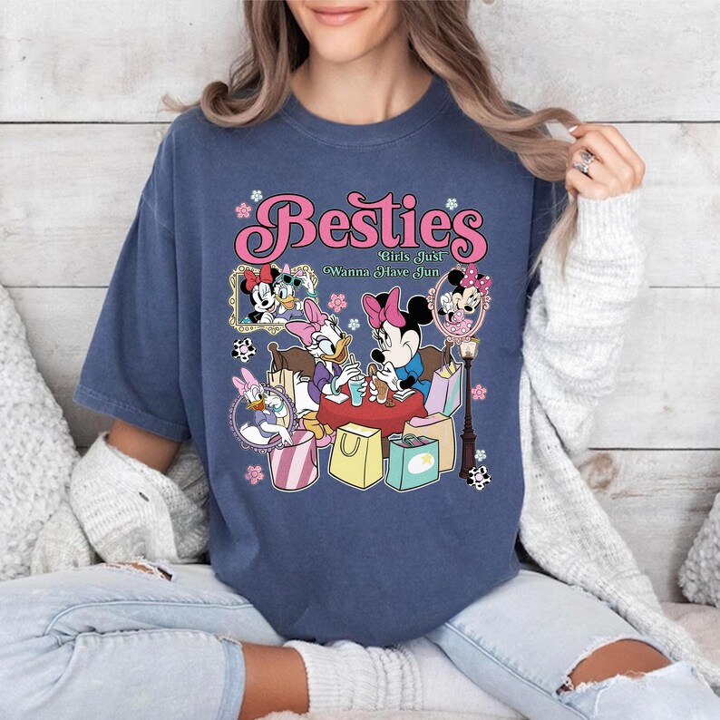 May include: A navy blue t-shirt featuring a graphic of Minnie Mouse, Daisy Duck, and other characters. The text "Besties Girls Just Wanna Have Fun" is displayed in pink. The design includes shopping bags and floral accents.