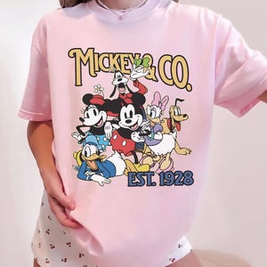 May include: Pink t-shirt featuring a graphic of Mickey Mouse and friends, with the text "Mickey & Co. EST. 1928" in a vintage style. The design includes Mickey, Minnie, Donald Duck, Goofy, Daisy, and Pluto.