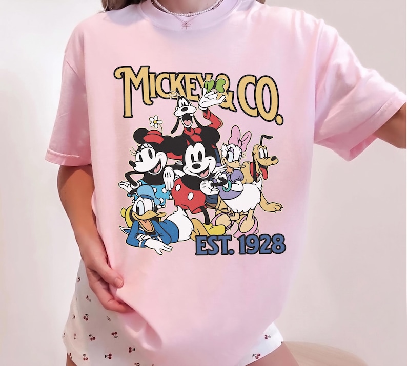 May include: Pink t-shirt featuring a graphic of Mickey Mouse and friends, with the text "Mickey & Co. EST. 1928" in a vintage style. The design includes Mickey, Minnie, Donald Duck, Goofy, Daisy, and Pluto.