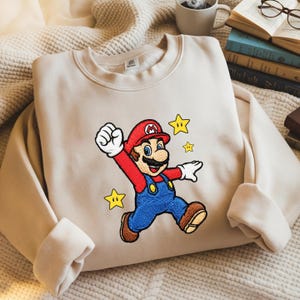 May include: Beige sweatshirt featuring an embroidered image of Mario, a popular video game character. Mario is depicted in his signature red hat, blue overalls, and brown shoes, with yellow stars around him. The sweatshirt is a neutral color.