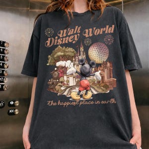 May include: Dark gray t-shirt featuring a vintage-style graphic of Mickey Mouse in front of a Disney World scene. The text "Walt Disney World" and "The happiest place in earth" are printed above and below the graphic.